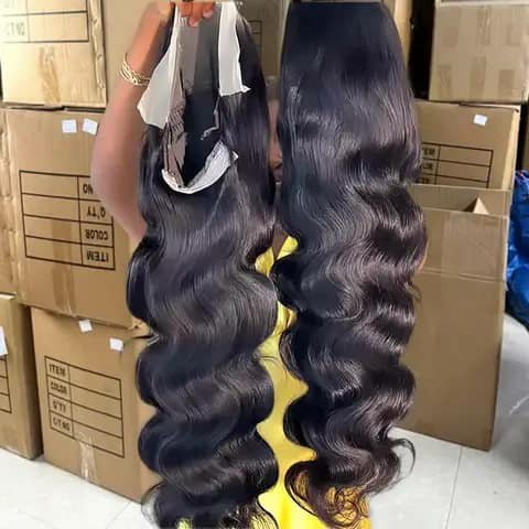 A person holding up two long black body-wave wigs in a warehouse.