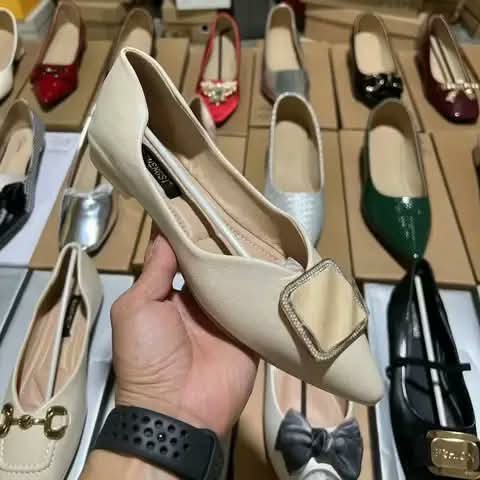 A cream flat shoe with a square buckle, held in hand.