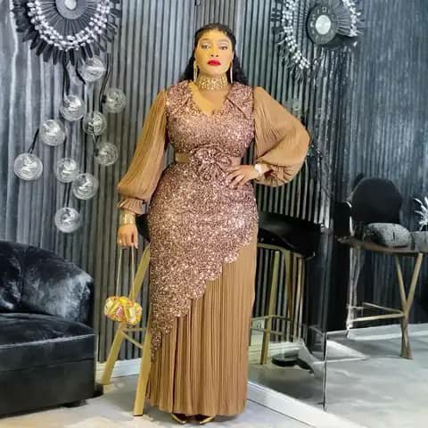 A woman wearing a long bronze sequinned dress with a pleated skirt and puffed sleeves, holding a bag, indoors.