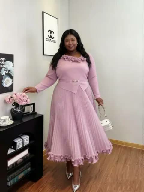A woman wearing a long light-pink pleated dress with a ruffled neckline, holding a white handbag, indoors.