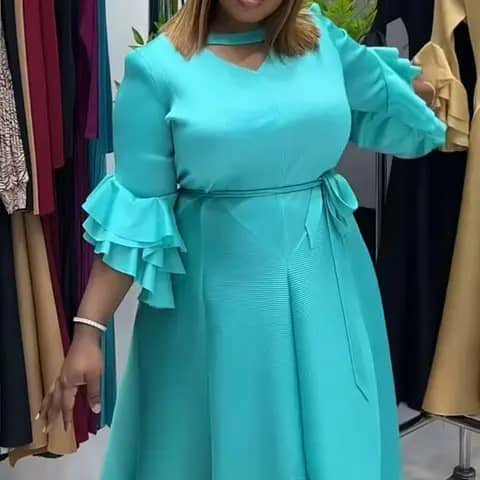 A woman wearing a turquoise dress with ruffled sleeves, a choker neckline and a waist tie, indoors.