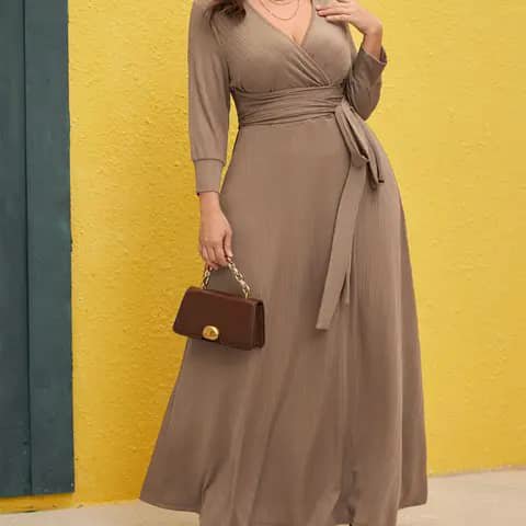 A woman wearing a taupe wrap maxi dress with a waist sash, holding a small brown handbag.