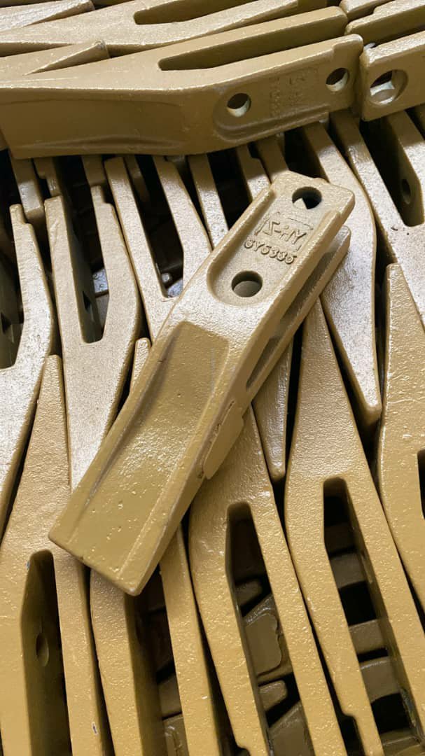 A pile of tan metal excavator bucket teeth, one stamped with markings.
