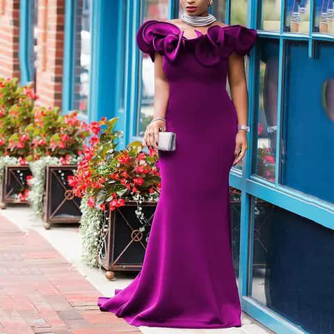 A woman wearing a long purple satin gown with a ruffled off-shoulder neckline, holding a clutch, outdoors.