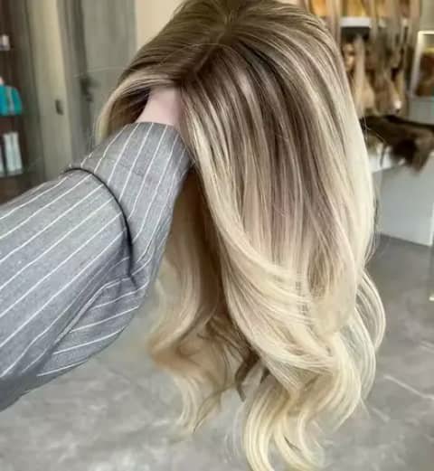 Long blonde wavy wig with dark roots, held up in a salon